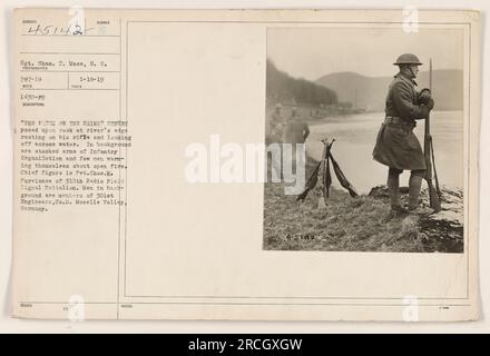 Chap. Pvt. H. Purviance du 310th radio Field signal Battalion se pose comme une sentinelle sur un rocher au bord de la rivière, regardant à travers l'eau. En arrière-plan, les membres du 301st Engineers, Co. D peut être vu se réchauffer autour d'un feu ouvert, avec des armes empilées d'une organisation d'infanterie visibles. La photographie a été prise dans la vallée de la Moselle, en Allemagne. (Crédit photo : Sgt. Shas. F. Mace, S.C.) Banque D'Images