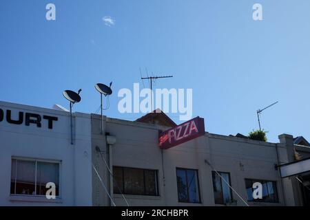 Un panneau de pizza rose et rouge au néon brille dans la lumière de l'après-midi, deux antennes paraboliques et des antennes de télévision montées sur le toit au-dessus d'une rangée de magasins Banque D'Images