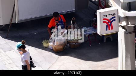 Vue aérienne : un vendeur de nourriture de rue, un homme, est assis sur le trottoir dans le centre de Bangkok, Thaïlande, Asie, attendant les clients. Une femme passe devant. L'emplacement est à côté de la station Siam BTS ou Skytrain. Affiche de rentrée des classes en haut à droite. Banque D'Images