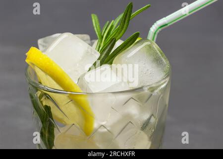 Morceaux de glace, quartiers de citron et branche de romarin dans le verre. Partie du cadre. Gros plan. Fond gris. Banque D'Images