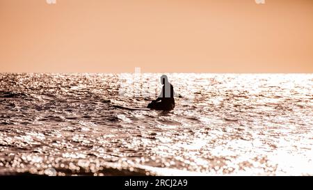 Silhouette de personne assise sur une planche de surf sur l'océan. Ciel dégagé, attendant une vague Banque D'Images