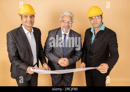 Jeunes ingénieurs indiens ou architectes portant un costume formel noir et un casque jaune ou un casque tenant le plan isolé sur fond beige. CONSTRU Banque D'Images
