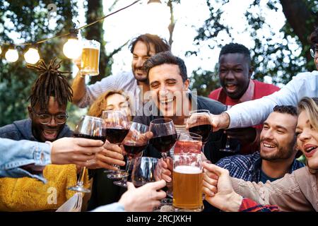 Un groupe diversifié d'amis âgés de 20 à 40 ans, grillant avec de la bière, du vin et des verres de sangria à l'extérieur dans une maison de campagne au crépuscule Banque D'Images