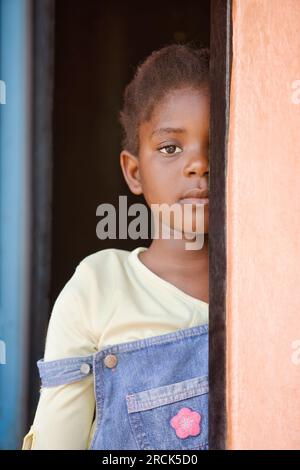 timide petite fille africaine avec des tresses se cachant dans le cadre de la porte à sa maison dans le township Banque D'Images