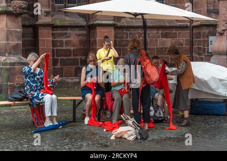 Kaiserslautern, Allemagne. 15 juillet 2023. Frau Elfride Peil et les spectateurs femmes tricotent de longues écharpes rouges. Trois jours de spectacles de rue théâtraux et de musique offerts par 200 artistes internationaux dans le centre-ville de Kaiserslautern - jour 2. Crédit : Gustav Zygmund/Alamy News Banque D'Images