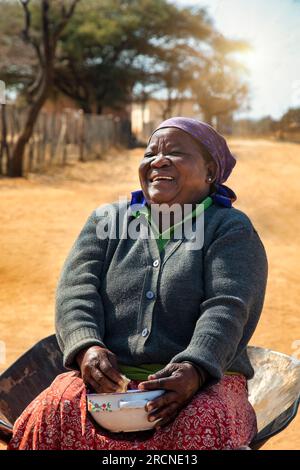 vie de village, vieille femme africaine située dans une brouette mangeant du ragoût et riant du cœur Banque D'Images