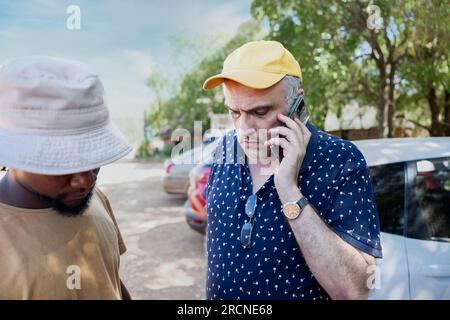 vieil homme avec un chapeau jaune, parlant au téléphone, son jeune ami africain à proximité, debout sur une route très fréquentée devant quelques voitures Banque D'Images