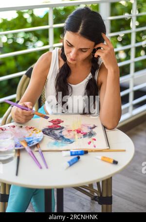 Femme brunette ukrainienne peignant le dessin à l'aquarelle d'une fille asiatique avec un pinceau sur une table avec de l'eau et des peintures Banque D'Images