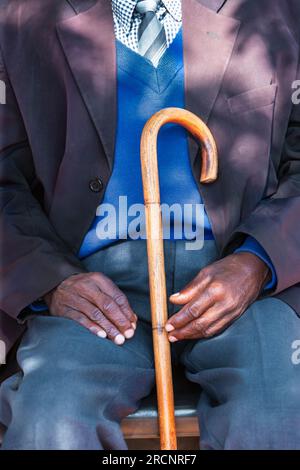 vieil homme africain fatigué et élégant avec un bâton de marche reposant sur une chaise. dehors dans la cour de son village Banque D'Images