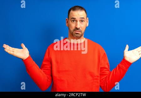 Homme hispanique barbu dans ses années 40 portant un sauteur rouge levant ses paumes dans un geste d'indifférence, isolé sur fond bleu de studio. Banque D'Images