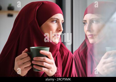 Femme musulmane dans le hijab avec une tasse de café regardant par une fenêtre. Banque D'Images