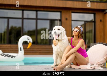 Femme embrasse avec son chien blanc mignon tout en se relaxant près de la maison avec une piscine. Amitié avec les animaux de compagnie et concept de vacances d'été Banque D'Images
