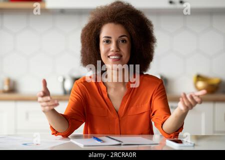 Souriant jeune femme afro-américaine parlant à la caméra tout en étant assise dans la cuisine Banque D'Images