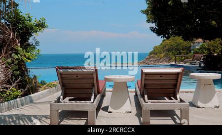 Chaises de plage en bois et une table près d'une piscine bleu clair avec vue sur la mer. Banque D'Images