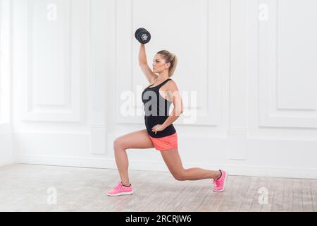 Portrait vue de côté de jeune athlétique belle femme blonde portant des shorts roses et dessus noir tenant haltère, faisant de l'exercice pour les bras à la salle de gym sur le mur blanc. Prise de vue en studio en intérieur. Banque D'Images