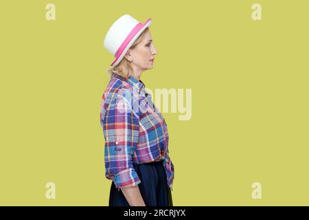 Portrait vue de côté de la femme mature sévère sérieuse portant chemise à carreaux et chapeau debout regardant devant, étant de mauvaise humeur. Studio intérieur tourné isolé sur fond jaune. Banque D'Images