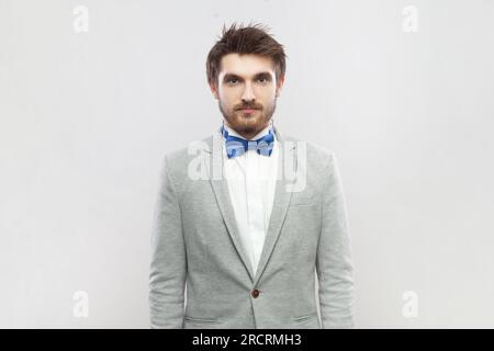 Portrait d'un homme barbu positif et calme se tenant debout devant la caméra, ayant une expression faciale détendue, portant un costume gris et un noeud papillon bleu. Studio intérieur tourné isolé sur fond gris. Banque D'Images