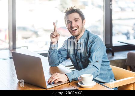 Portrait de jeune homme freelance excité et joyé en chemise bleu jeans travaillant sur ordinateur portable et levé le doigt, ayant une bonne idée, regardant la caméra. Prise de vue intérieure près de la grande fenêtre, fond de café. Banque D'Images