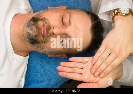 Massothérapeute faisant un massage facial pour homme dans le salon de spa. Bel homme barbu recevant un traitement de soin de la peau de l'esthéticienne. Modelage facial actif Banque D'Images