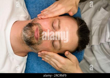 Massothérapeute faisant un massage facial pour homme dans le salon de spa. Bel homme barbu recevant un traitement de soin de la peau de l'esthéticienne. Modelage facial actif Banque D'Images