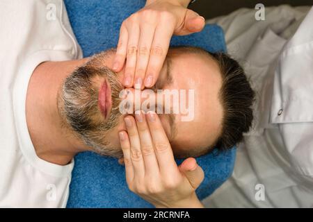 Masseuse faisant massage facial pour homme dans le salon de spa. Bel homme barbu recevant un traitement de soins de la peau de cosméticien. Modelage actif du massage facial Banque D'Images