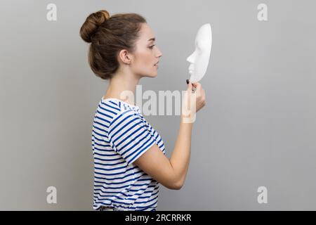 Portrait de vue latérale de belle femme aux cheveux sombres avec coiffure de pain portant T-shirt rayé debout tenant et regardant le masque blanc. Studio intérieur tourné isolé sur fond gris. Banque D'Images