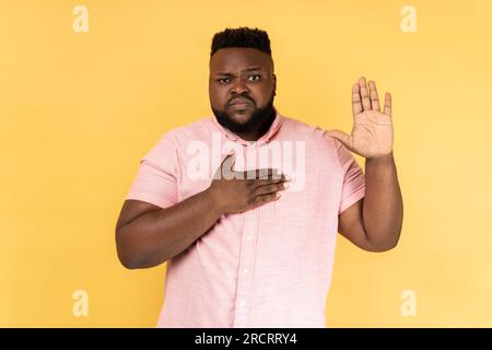 Portrait d'homme patriotique sérieux portant une chemise rose tenant la main sur le cœur, juré jurant de dire la vérité au tribunal, l'honneur et la conscience. Studio intérieur tourné isolé sur fond jaune. Banque D'Images