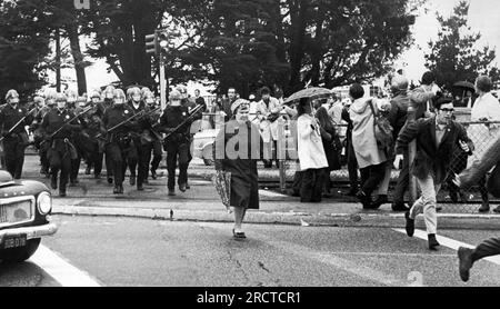 San Francisco, Californie : 9 décembre 1968 une dame de parapluie qui traverse la 19e Avenue, ignorant complètement que l'escadron de police en équipement anti-émeute double chronométrage derrière elle à la poursuite des manifestants fuyant l'Université d'État de San Francisco. Banque D'Images