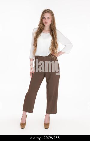 Jeune femme blonde aux cheveux longs portant un chemisier blanc, un pantalon marron et des chaussures debout en pleine longueur, poses. Femme d'affaires caucasienne 21 ans Banque D'Images