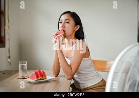 Une belle femme asiatique se sentant mal à l'aise par le temps chaud, mange de la pastèque et s'assoit devant un ventilateur électrique pour se rafraîchir à la maison dans TH Banque D'Images