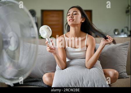 Une femme asiatique fatiguée et surchauffée souffrant d'une attaque de chaleur, est assise sur un canapé devant un ventilateur électrique et utilise un ventilateur pratique pour se rafraîchir Banque D'Images