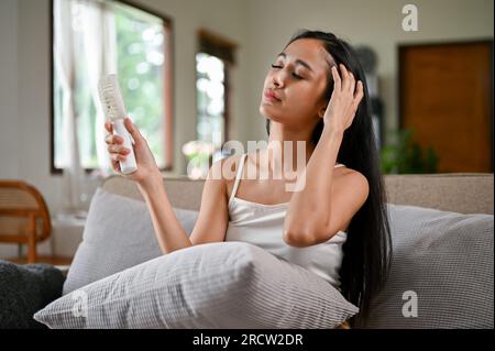 Une femme asiatique surchauffée se sentant inconfortable suite à une attaque de chaleur, utilise un ventilateur électrique pratique pour se rafraîchir tout en se reposant sur un canapé dans son vivant Banque D'Images