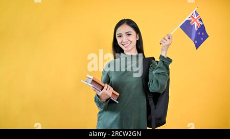 Une jolie étudiante asiatique en vêtements décontractés tient ses livres et le drapeau australien, debout sur un fond jaune et isolé. e Banque D'Images