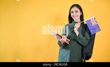 Une jolie étudiante asiatique dans des vêtements décontractés tient ses livres et le drapeau australien, debout sur un fond jaune, isolé wi Banque D'Images