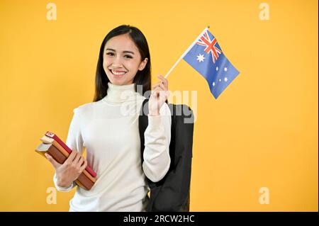 Une jolie étudiante asiatique tient ses livres et le drapeau australien, debout sur un fond jaune et isolé. Échangez l'étudiant an Banque D'Images