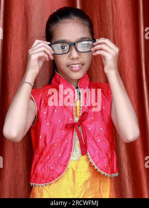 Une petite jolie fille indienne violonant avec ses lunettes dans diverses poses en robe rouge. Banque D'Images