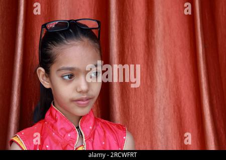 Une petite jolie fille indienne violonant avec ses lunettes dans diverses poses en robe rouge. Banque D'Images