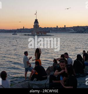 Les gens s'assoient sur des rochers en admirant le coucher de soleil sur la mer du Bosphore et la Tour Maidens d'Uskudar, Istanbul, Turquie. Tour de Galata et bateau de croisière derrière. Banque D'Images