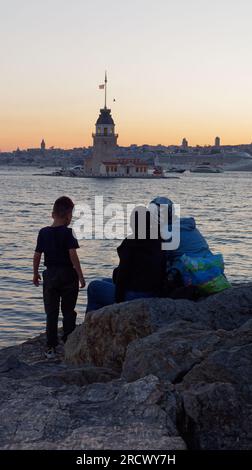 Les gens s'assoient sur des rochers en admirant le coucher de soleil sur la mer du Bosphore et la Tour Maidens d'Uskudar, Istanbul, Turquie. Tour de Galata et bateau de croisière derrière. Banque D'Images