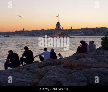 Les gens s'assoient sur des rochers en admirant le coucher de soleil sur la mer du Bosphore et la Tour Maidens d'Uskudar, Istanbul, Turquie. Tour de Galata et bateau de croisière derrière. Banque D'Images