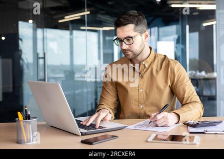 Sérieux jeune homme comptable, auditeur, analyste travaille dans le bureau à la table derrière l'ordinateur portable et avec des documents. Rédige des factures, fait des analyses, remplit des rapports financiers et fiscaux. Banque D'Images