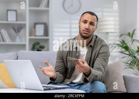 Portrait d'un jeune homme afro-américain contrarié regardant avec inquiétude la caméra et tenant une carte de crédit. Assis à la maison sur le canapé devant l'ordinateur portable et regardant avec déception la caméra. Banque D'Images