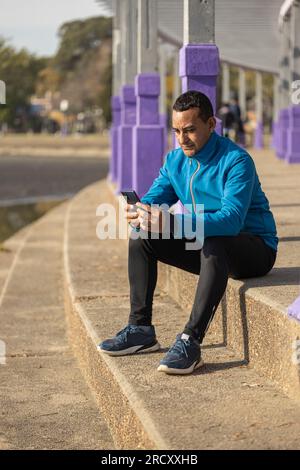 Jeune latino homme en vêtements de sport regarde son téléphone portable assis sur quelques escaliers. Banque D'Images