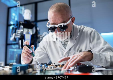 Programmeur mature en blouse de laboratoire assis à la table et assemblant la carte mère dans le bureau Banque D'Images