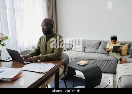 Père afro-américain assis à table et travaillant sur ordinateur portable à la maison pendant que son fils joue sur tablette pc en arrière-plan Banque D'Images