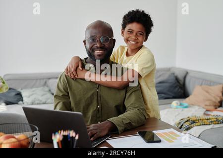 Portrait de fils heureux souriant à la caméra et embrassant son père pendant qu'il travaille sur ordinateur portable Banque D'Images