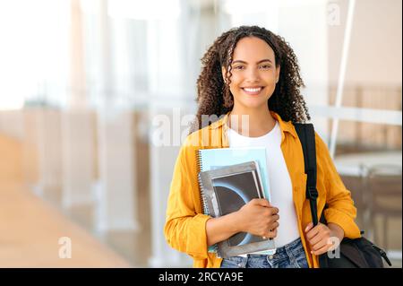 Heureuse belle étudiante brésilienne ou hispanique aux cheveux bouclés, avec un sac à dos, tenir des livres et des cahiers dans sa main, se tenir près du campus universitaire, regarder et sourire à la caméra. Copy-space Banque D'Images