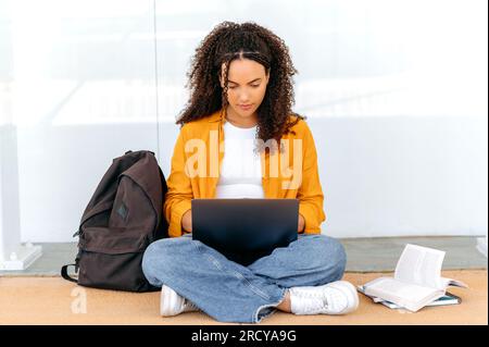 Belle étudiante brésilienne ou hispanique aux cheveux bouclés focalisée et élégante, asseyez-vous près du campus universitaire, en utilisant un ordinateur portable, se préparer à l'examen, faire ses devoirs, tâche de projet, sourire Banque D'Images