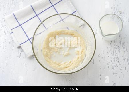 Bol en verre avec masse fouettée d'oeuf-crème-sucre, ajoutant du lait sur un fond bleu clair, vue de dessus. Cuisiner des gâteaux de lait faits maison, faites-le vous-même, étape par s. Banque D'Images