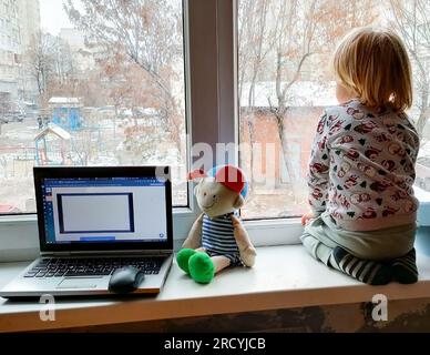 Un petit garçon aux cheveux blonds est assis près de la fenêtre sur le rebord de la fenêtre et regarde par la fenêtre. Hiver. Réveillon du nouvel an. L'enfant est habillé dans une nouvelle année' Banque D'Images